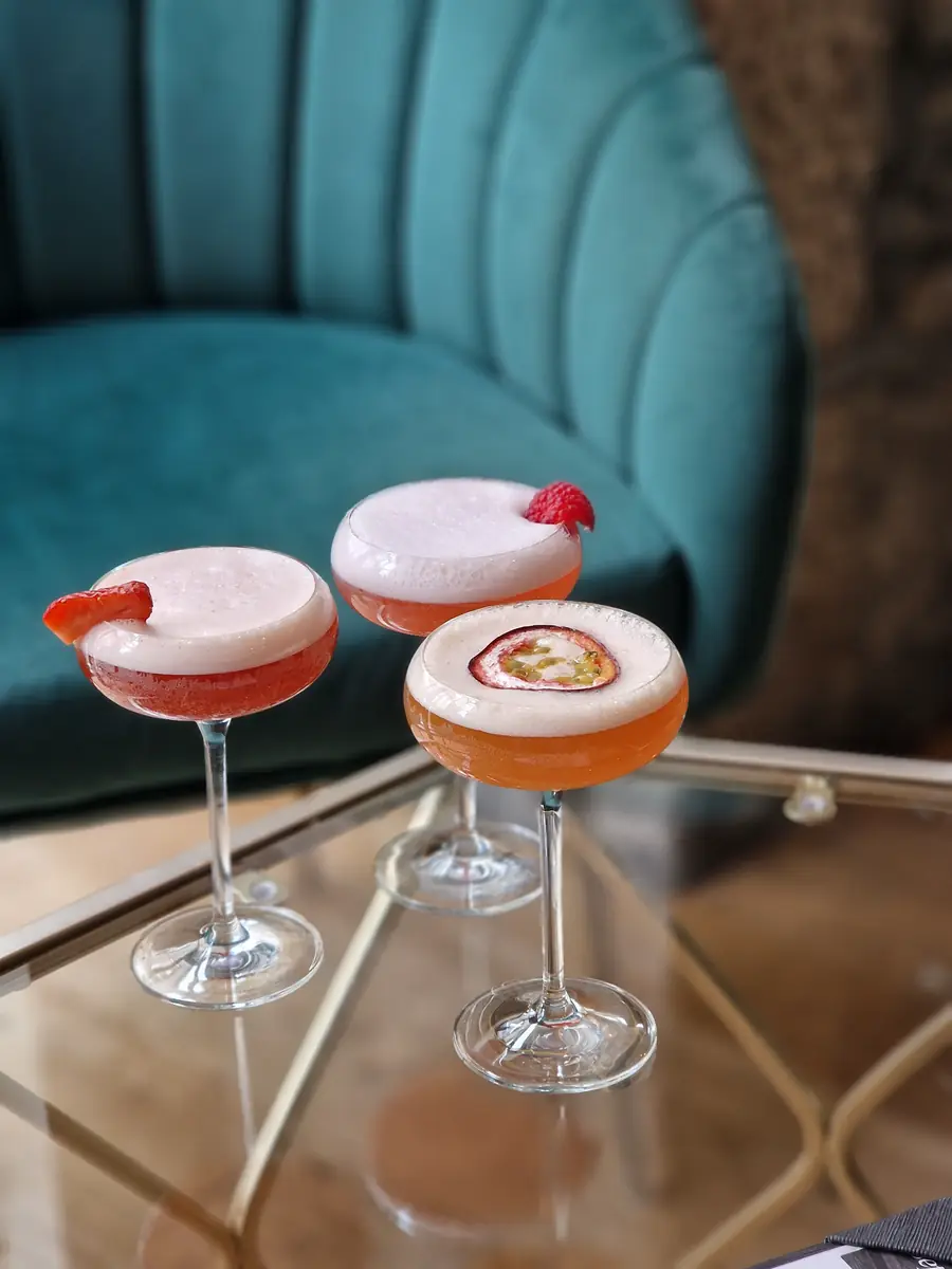 Three coupe glasses of cocktails garnished with strawberry, raspberry and passion fruit on a brass and glass table.