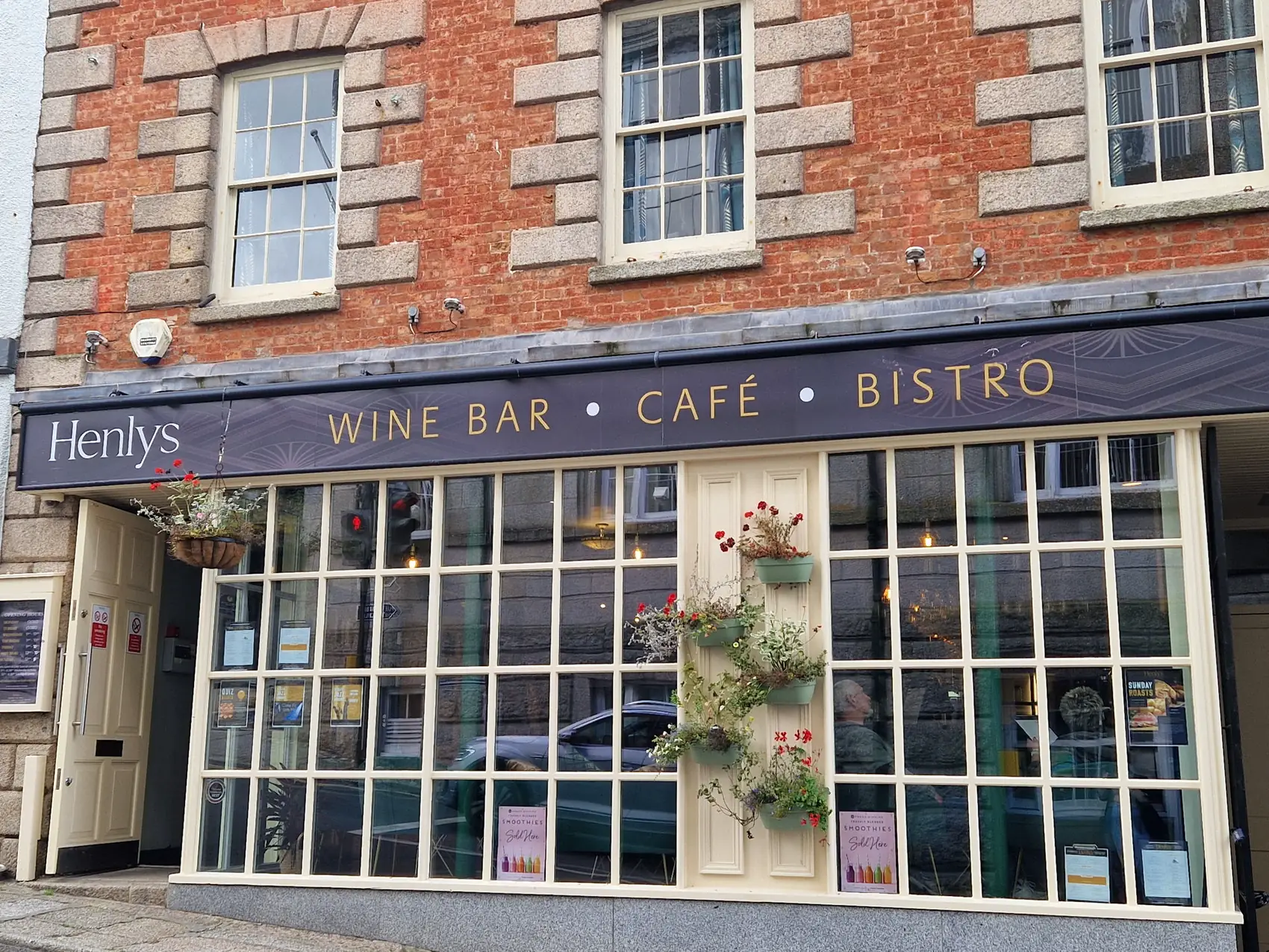 The Henlys shopfront on Church Street, Helston: a navy awning reading Wine Bar, Caf&eacute;, Bistro above tall Georgian windows and hanging baskets.